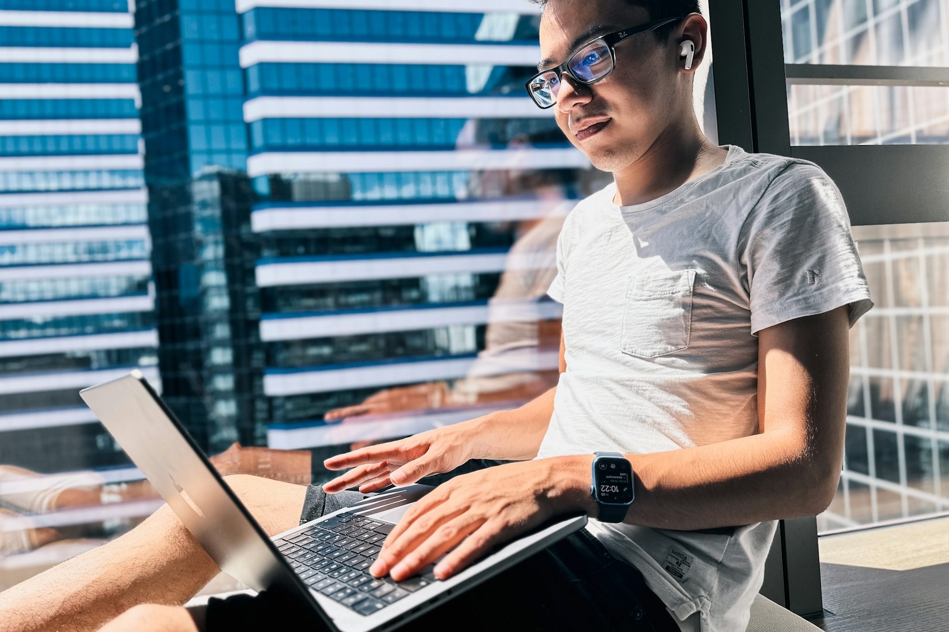 Young man working on a laptop by a window with city views.